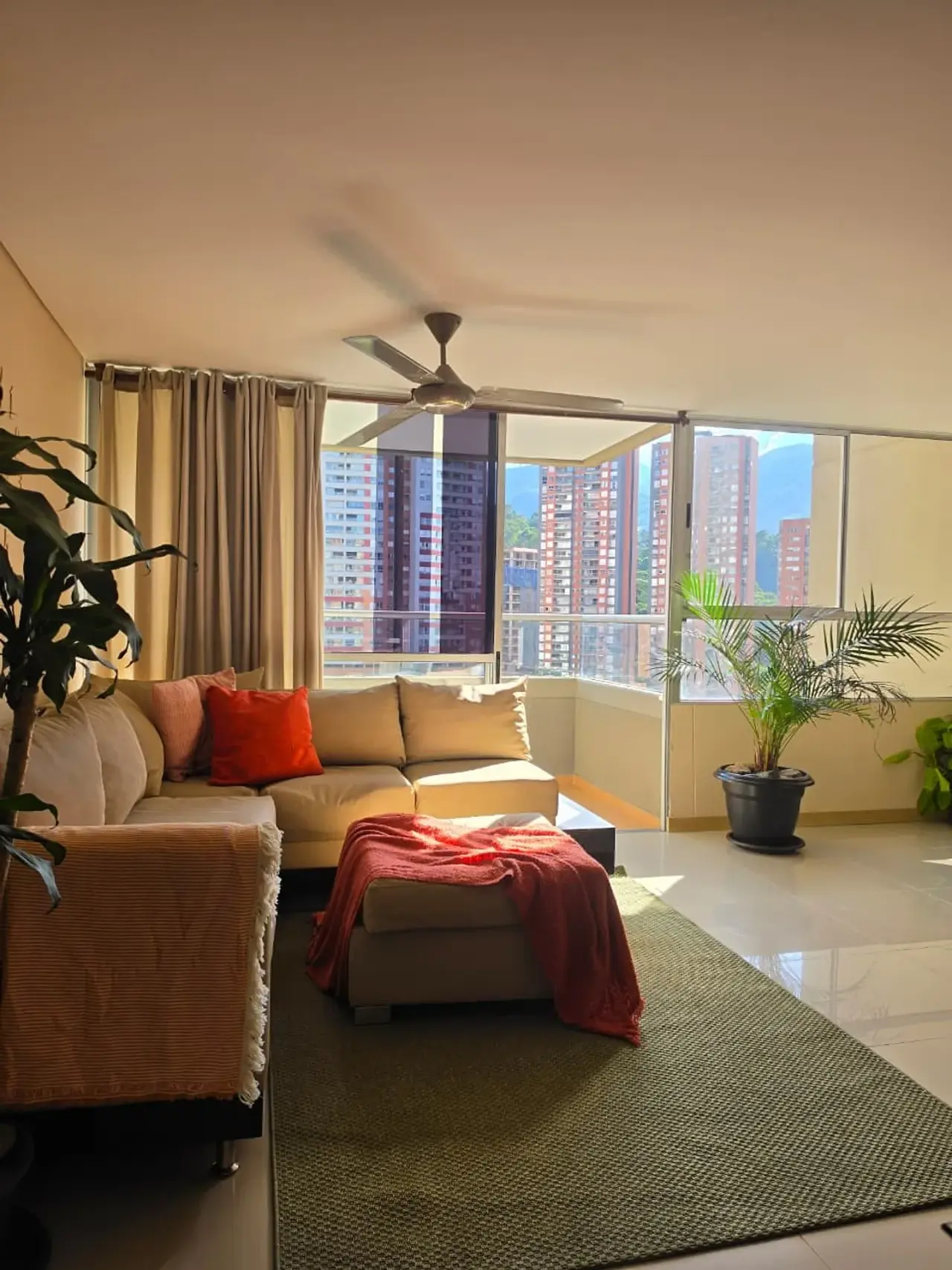Sabaneta Parkside — sunlit living room with a sectional sofa, plants and large windows onto Sabaneta's towers, Medellín
