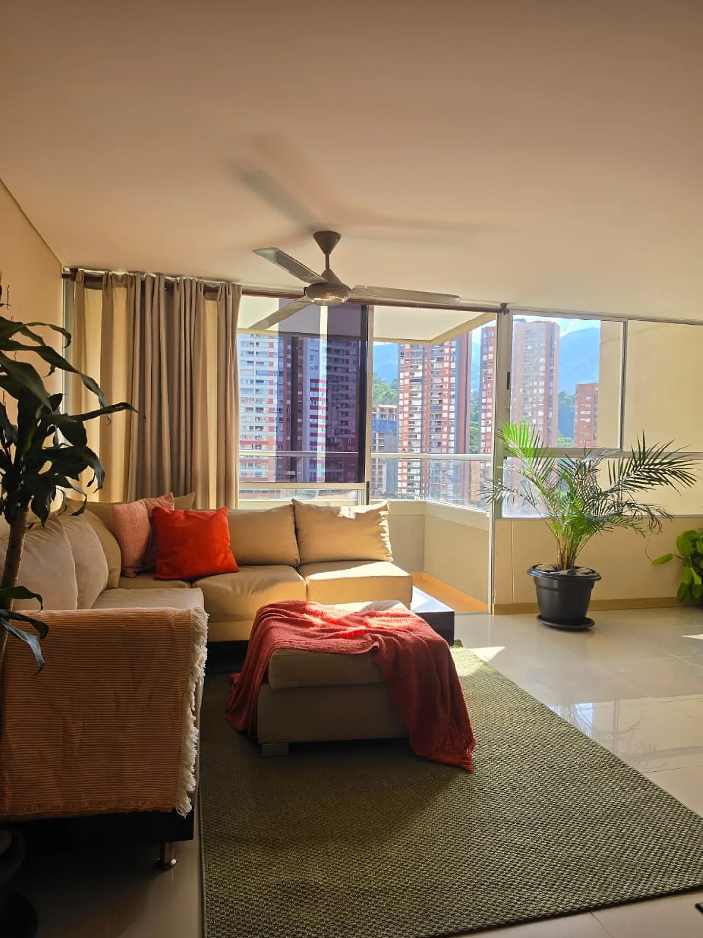 Sabaneta Parkside — sunlit living room with a sectional sofa, plants and large windows onto Sabaneta's towers, Medellín